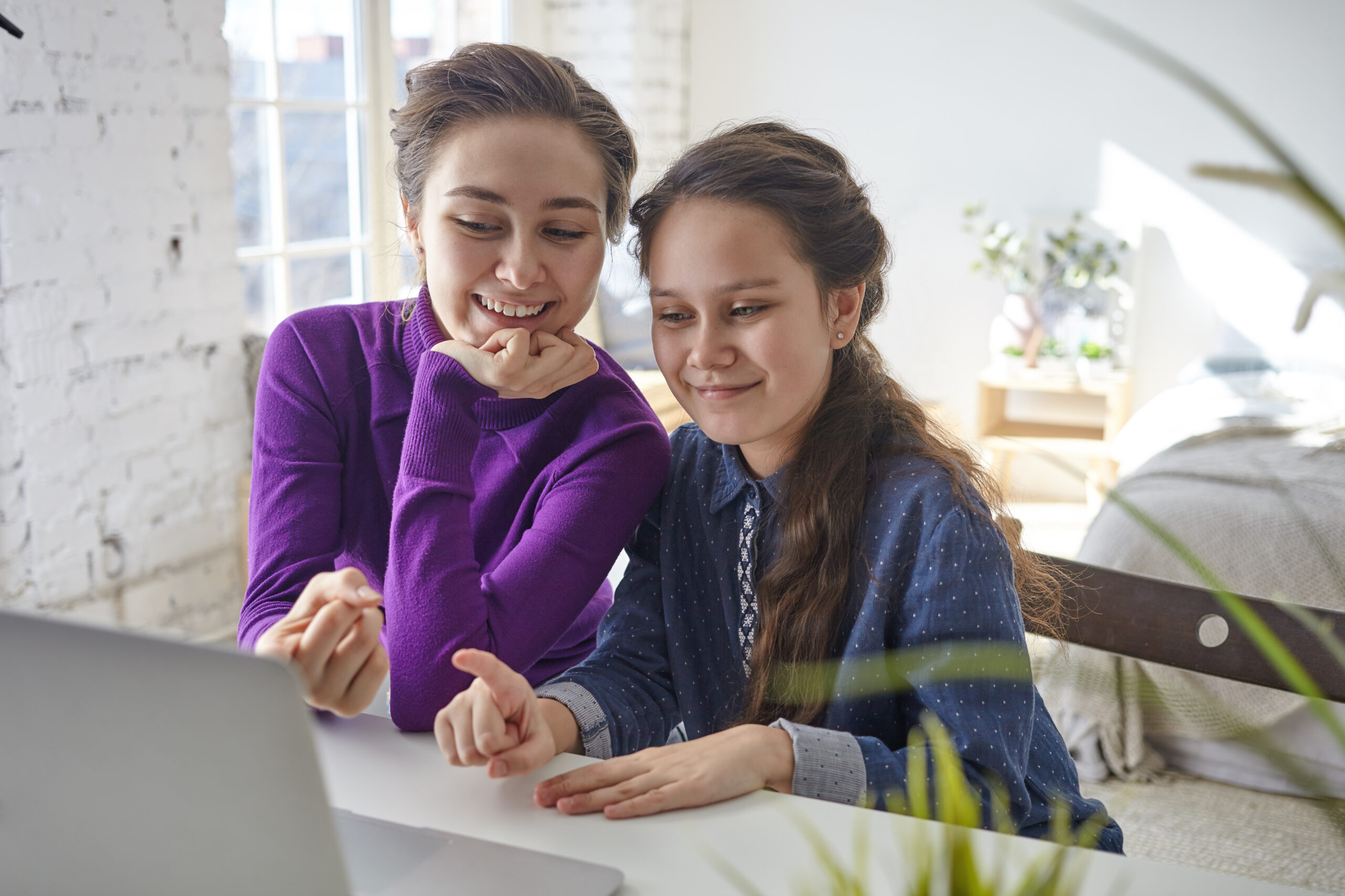 Candid shot of joyful happy young mother and daughter viewing family pictures or shopping online using laptop pc, sitting at desk in light bedroom interior, pointing fingers at screen and smiling Candid shot of joyful happy young mother and daughter viewing family pictures or shopping online using laptop pc, sitting at desk in light bedroom interior, pointing fingers at screen and smiling