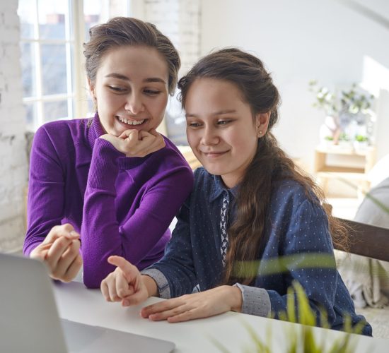 Candid shot of joyful happy young mother and daughter viewing family pictures or shopping online using laptop pc, sitting at desk in light bedroom interior, pointing fingers at screen and smiling